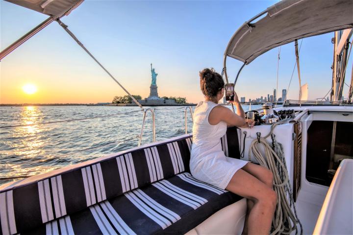 woman wearing a white dress and seated on a blue and white striped cushion taking a photo of the Statue of Liberty during sunset on a private boat cruise NYC