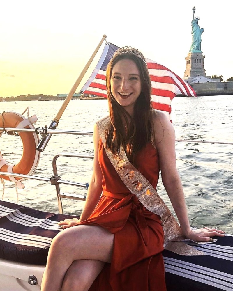 a woman sitting on a birthday boat in NYC with the Statue of Liberty behind her