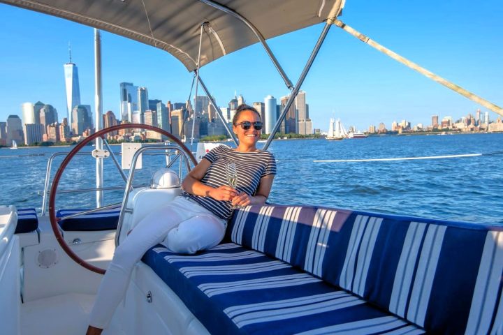 woman in a blue striped shirt sitting on a private boat charter nyc with a steering wheel and Manhattan skyline behind her