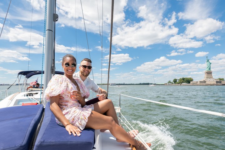 couple sitting on a blue cushion drinking champagne by the statue of liberty while on a new york sailboat rental