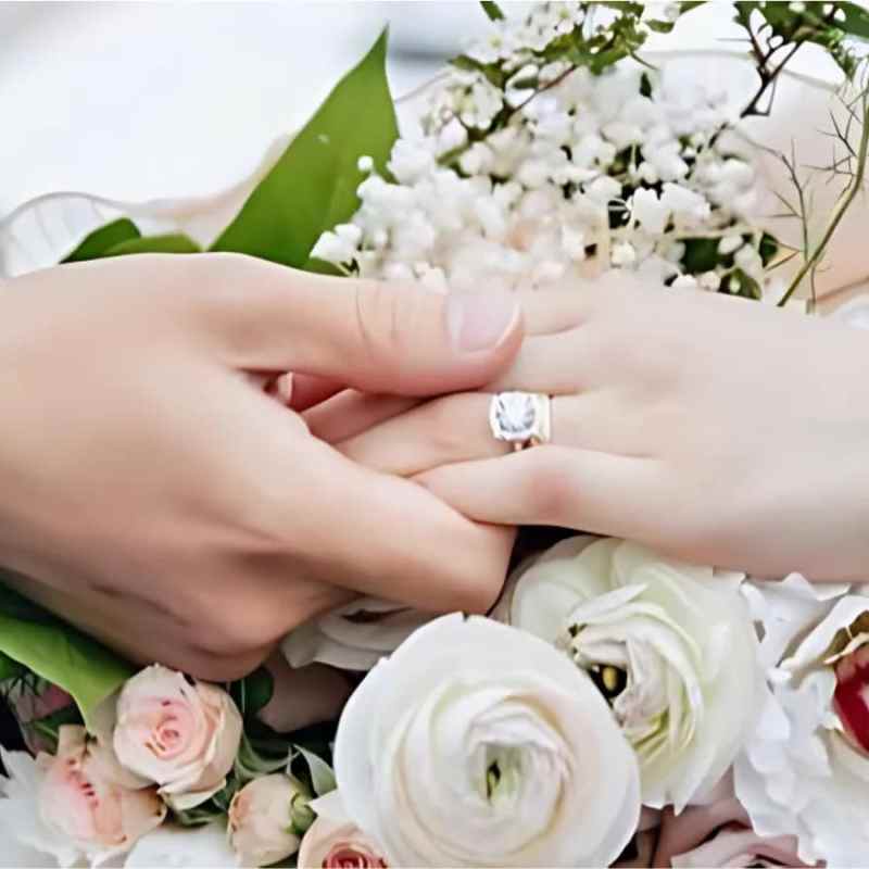 Two hands clasped over a bouquet of white and pink flowers, one wearing a ring and bracelet.