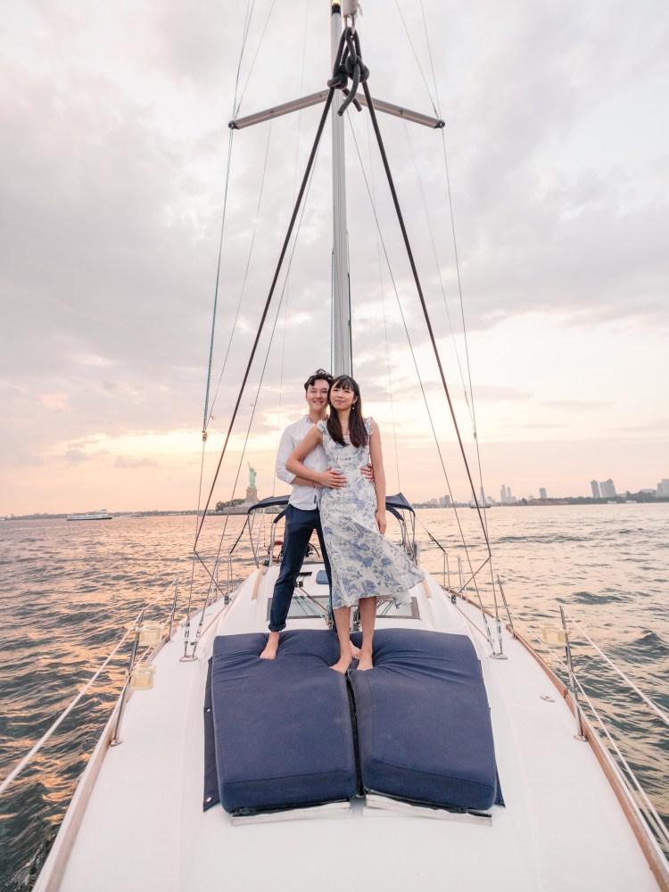 couple rent a sailing yacht and stand on blue deck cushions with the Statue of Liberty in the background