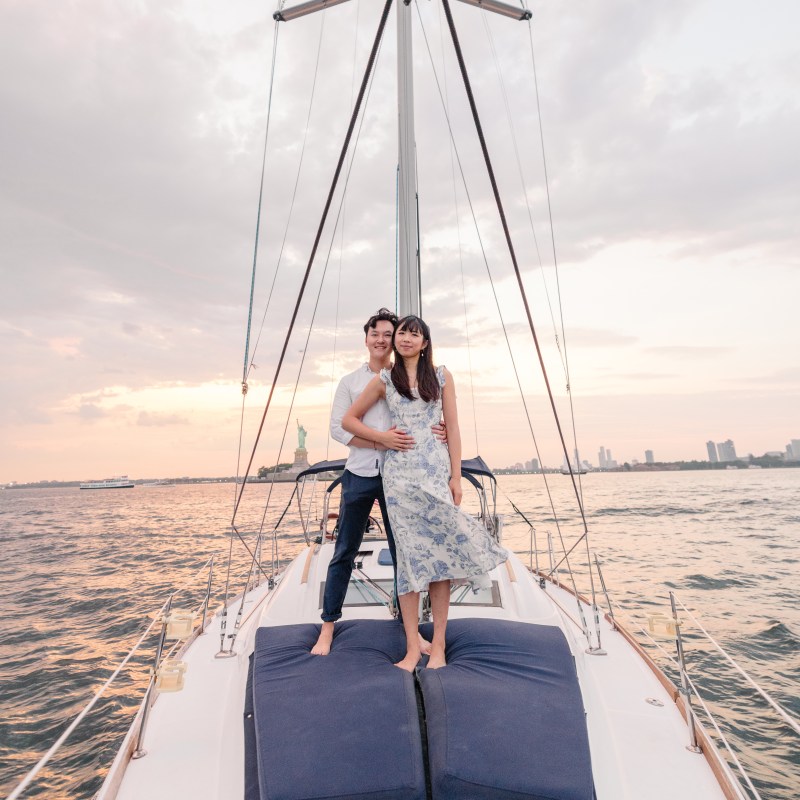 couple rent a sailing yacht and stand on blue deck cushions with the Statue of Liberty in the background