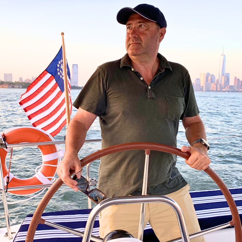 Captain Martin steering private boat rental through New York Harbor with a flag and city skyline in background at sunset.