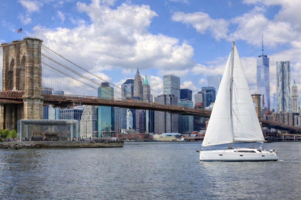 Sailboat on river with Brooklyn Bridge and New York City skyline in background.