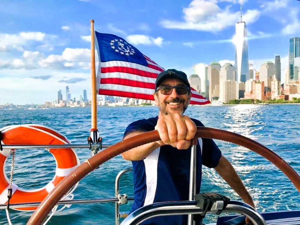 USCG licensed captain steering a sailboat charter NYC in New York Harbor with the skyline and US flag in background.