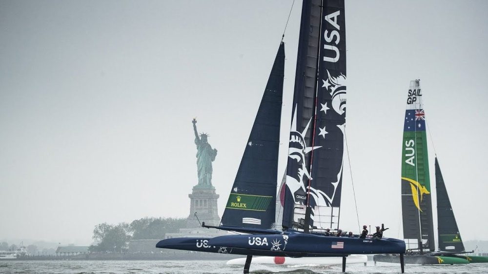 Sailboats with USA and AUS flags racing near Statue of Liberty from the perspective of a boat charter in NYC.