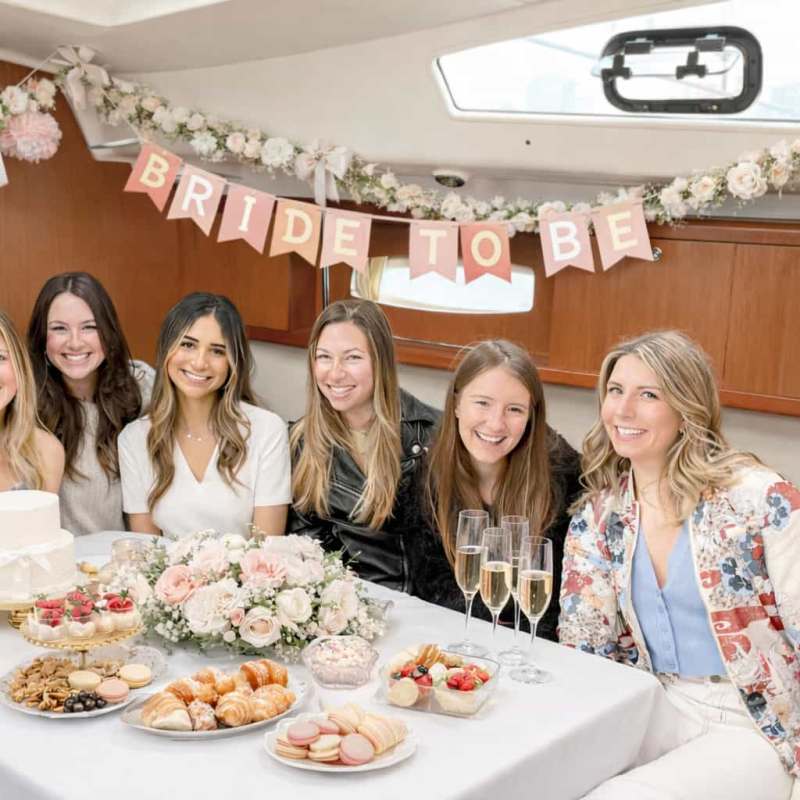 Six women at a table with desserts and 'Bride to Be' banner, smiling on a bachelorette party boat rental in NYC.