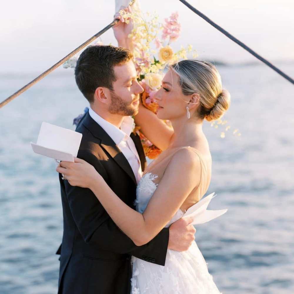 A couple in wedding attire embrace on a NYC boat charter, holding letters with flowers in the background.