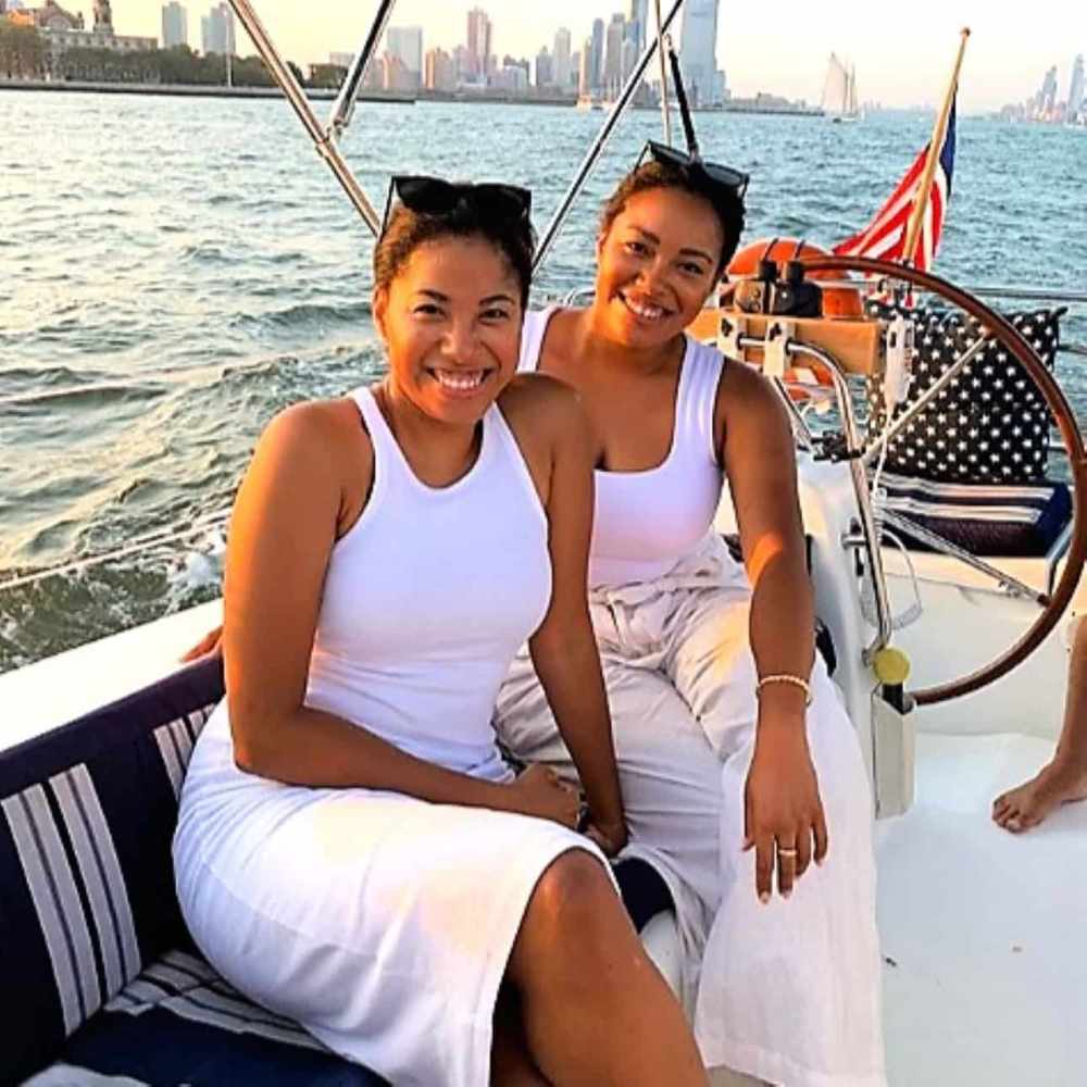 Two women in white outfits smiling on a party boat with the NYC skyline in the background.