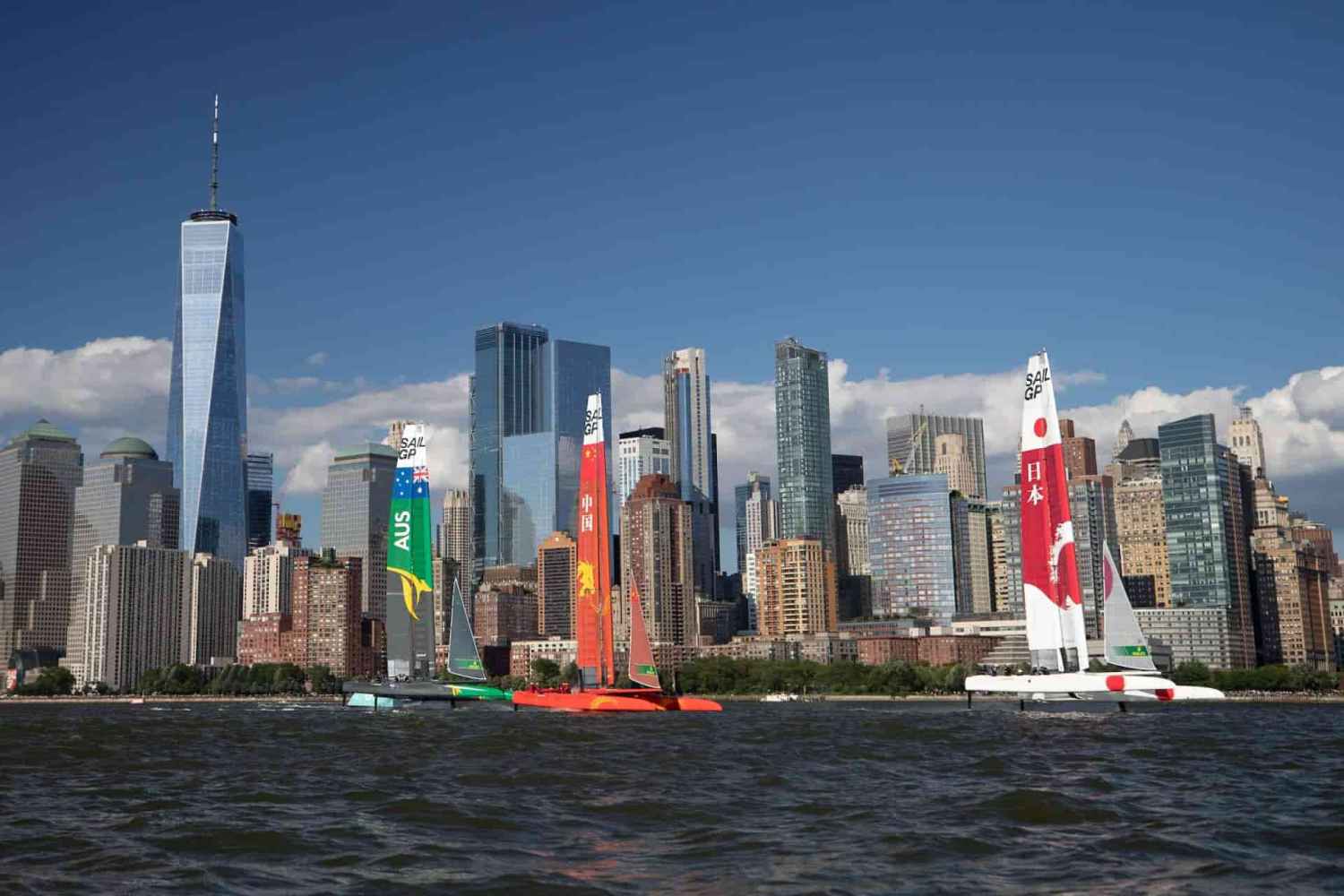 Colorful sailboats race during SailGP in front of the NYC skyline with a tall glass skyscraper during a private boat charter.