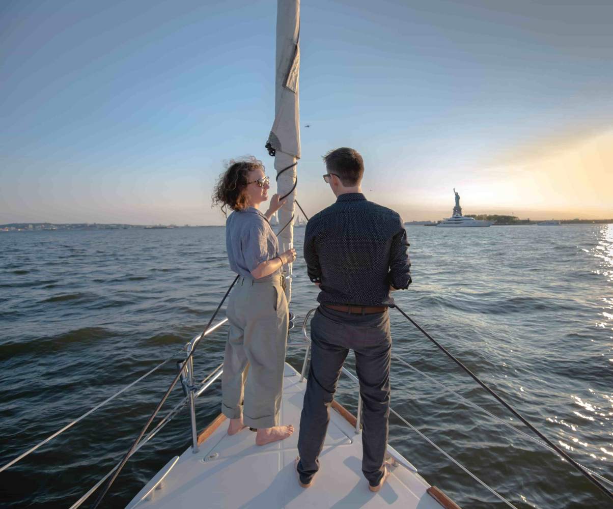 Two people on a private sailboat charter in NYC near the Statue of Liberty at sunset.