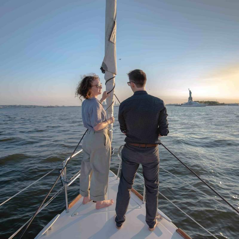 Two people on a private sailboat charter in NYC near the Statue of Liberty at sunset.