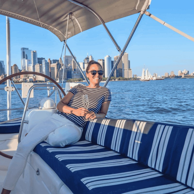 Person relaxing on a boat with a city skyline in the background under a clear blue sky.