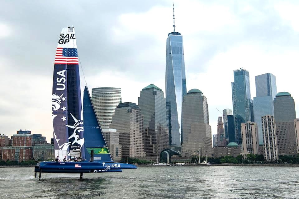 USA SailGP boat sailing past skyscrapers and waterfront buildings during a private charter in NYC.