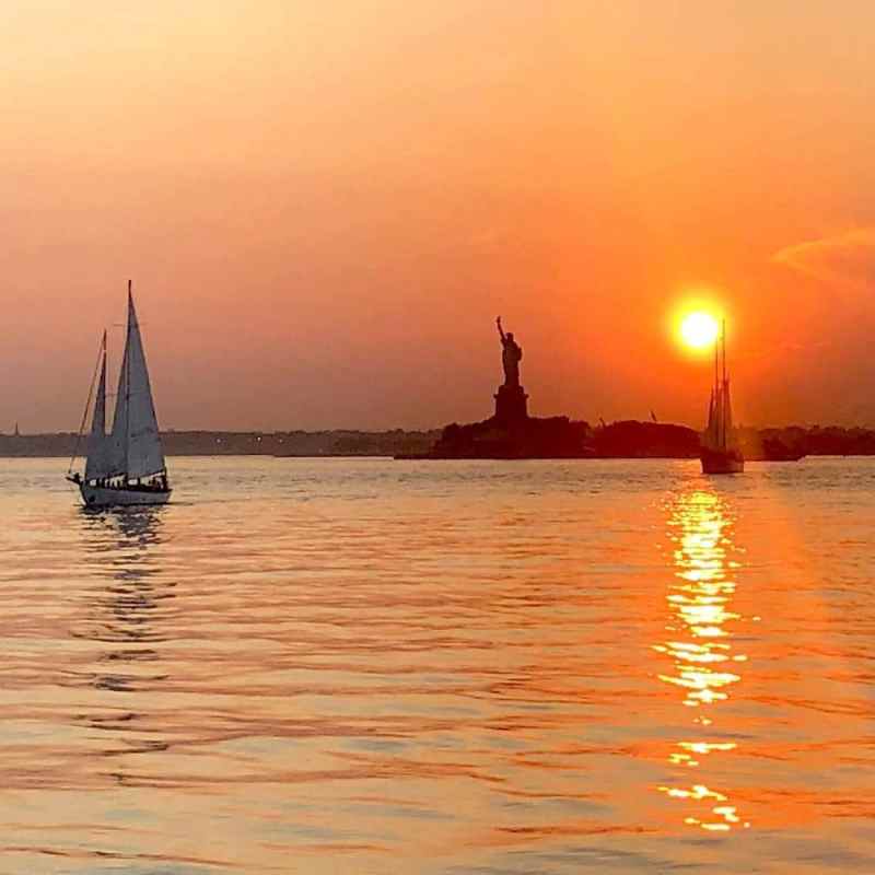 Sailboats near the Statue of Liberty at sunset, casting reflections on calm water during private cruises in NYC