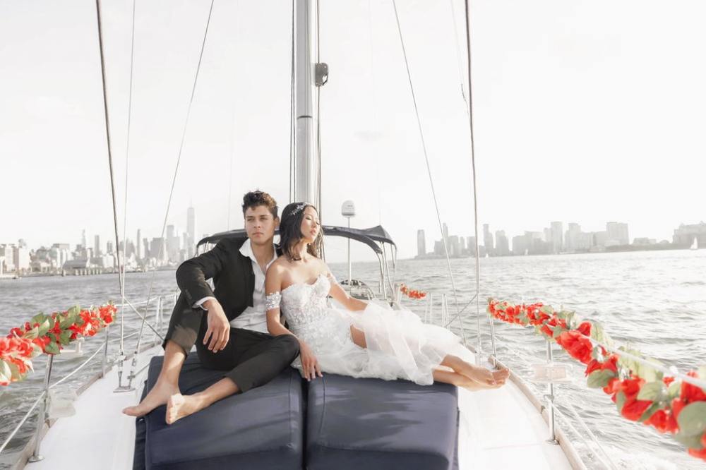 Couple in wedding attire on sailboat, NYC skyline in background.