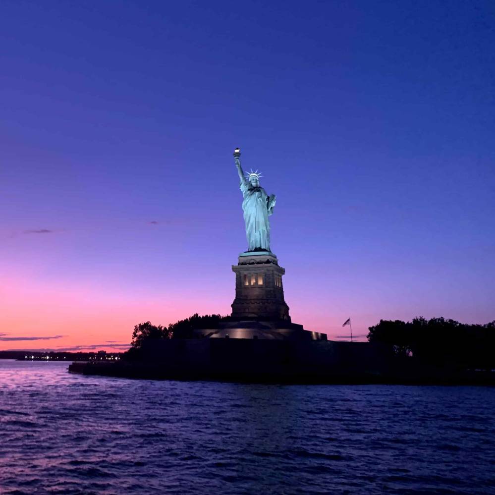 Statue of Liberty silhouetted against a colorful sunset sky over water during a sail in NYC.