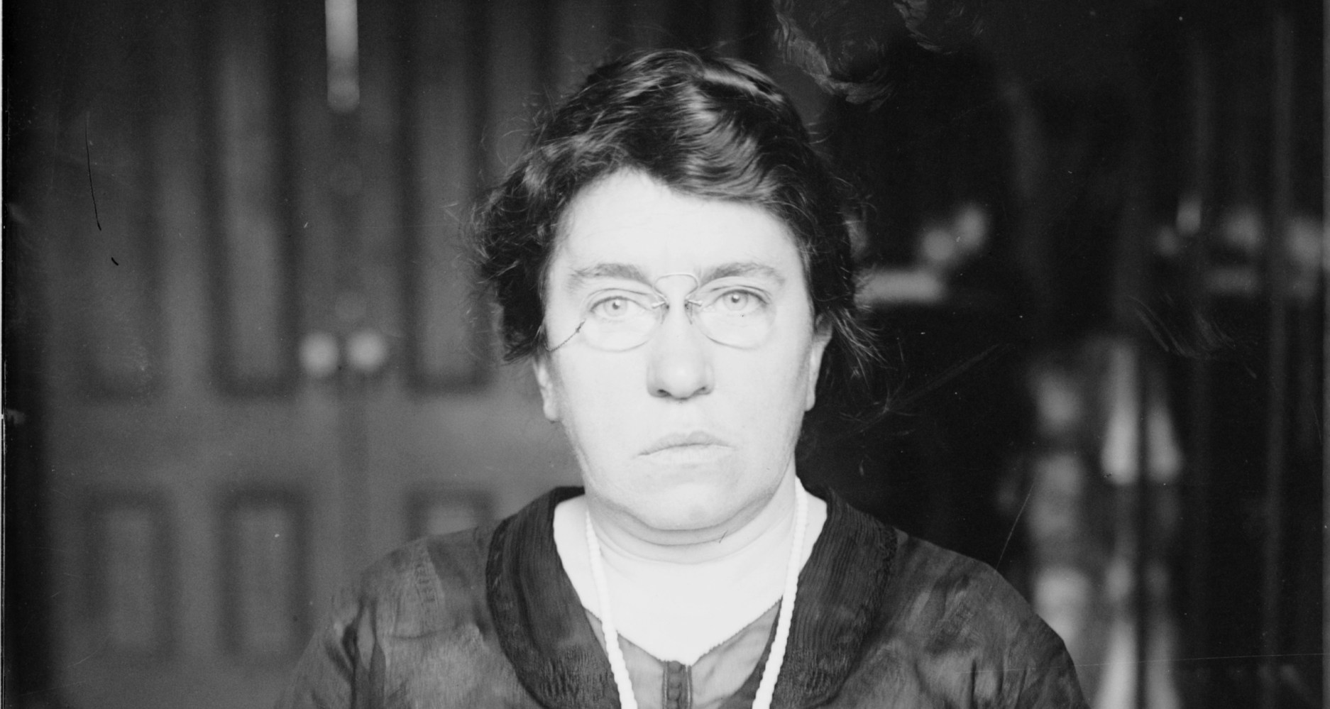Black and white portrait of a woman with glasses, wearing a dark top and facing forward.
