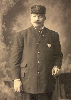 Vintage photo of man in uniform with a cap, standing beside chair.
