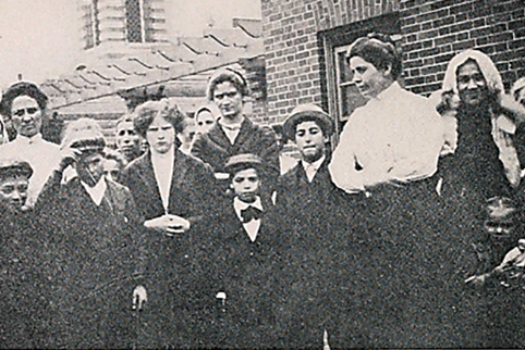 Vintage photo of a group of men and women posing outside near a brick building.