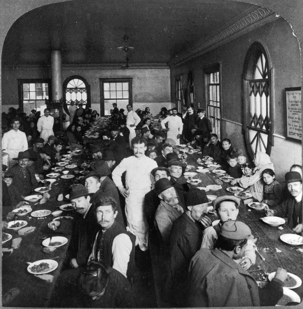 Historical photo of people dining in a large hall with long tables and benches.